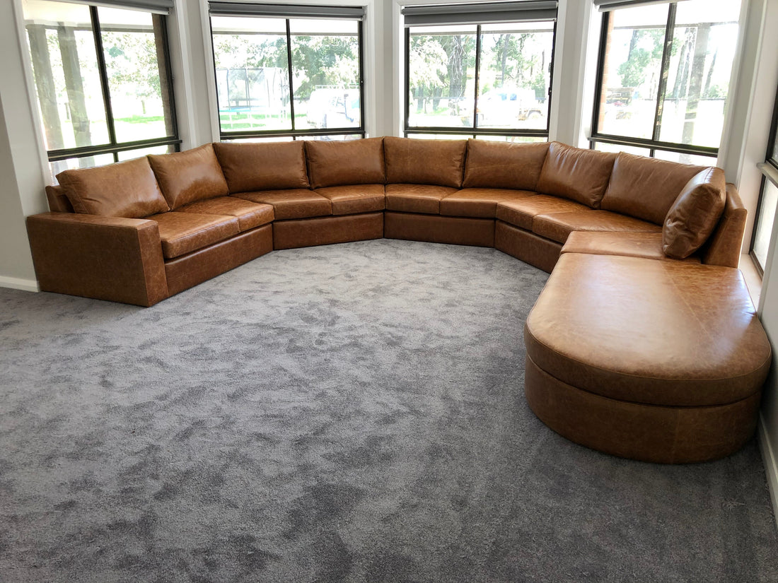 Custom caramel Italian aniline leather lounge wrapping around bay windows and angled walls in a country style living room with grey carpet.